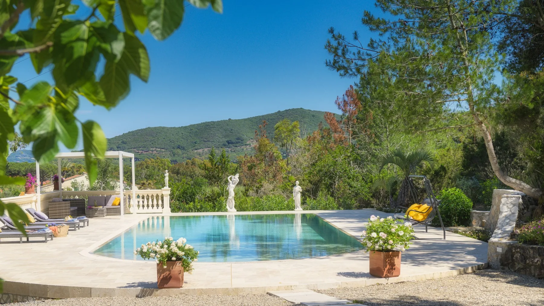 Una piscina está en una terraza soleada rodeada de árboles y vegetación.