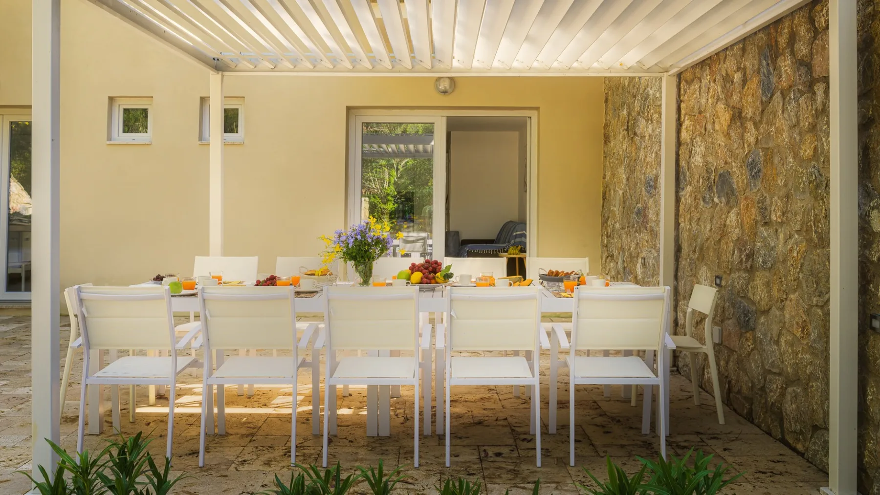 Comedor exterior cubierto con mesas y sillas blancas junto a una pared de piedra.