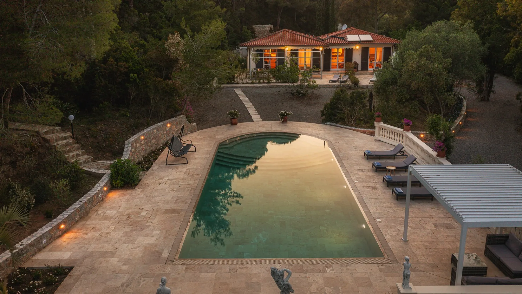 Una piscina y un patio pavimentado están frente a un edificio iluminado entre árboles.