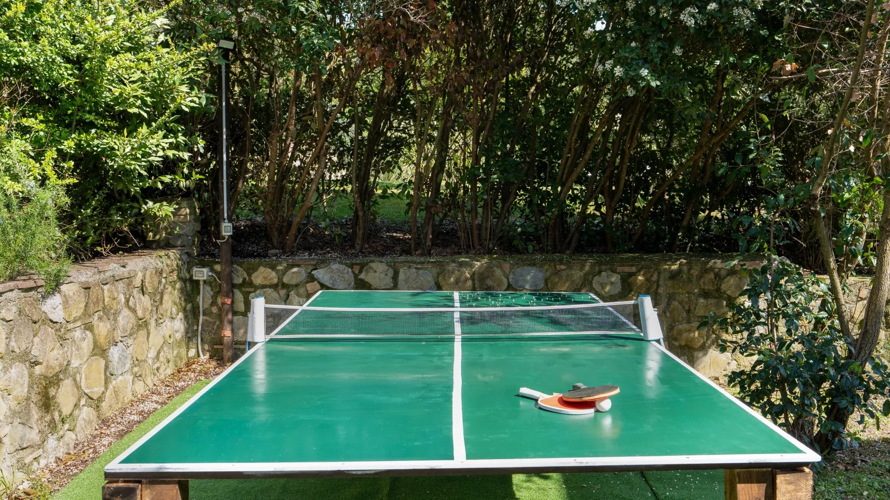 A green table tennis table stands outdoors beside a stone wall and trees.