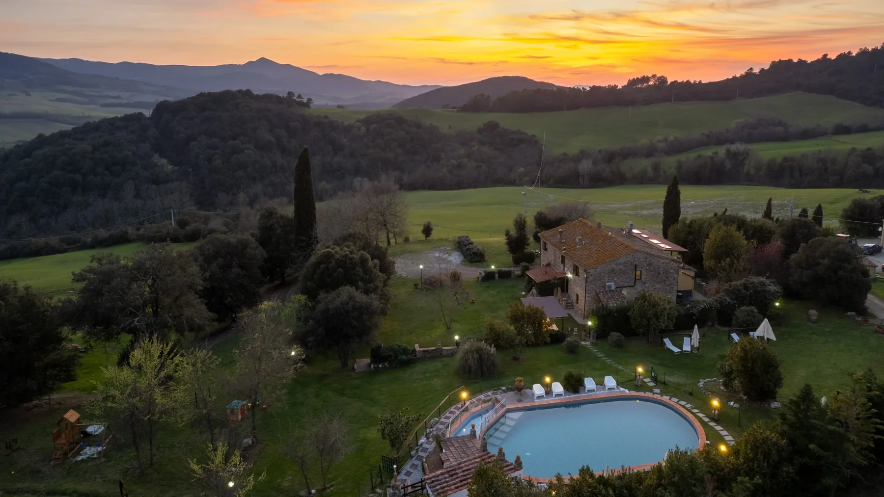 A countryside property with a swimming pool sits among rolling green hills at sunset.