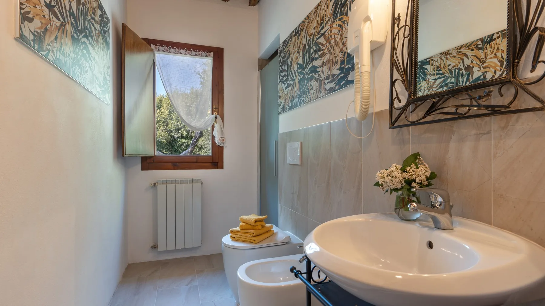 A bathroom with a sink, bidet, and window overlooking greenery.