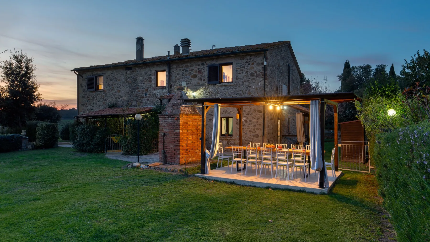 Stone house with a covered outdoor dining area beside the lawn at dusk.