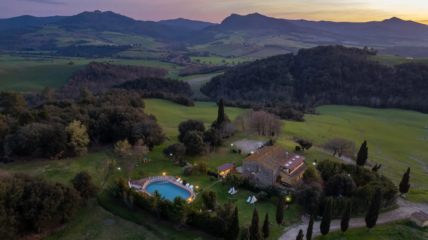 A countryside property with a swimming pool sits among green fields and wooded hills.