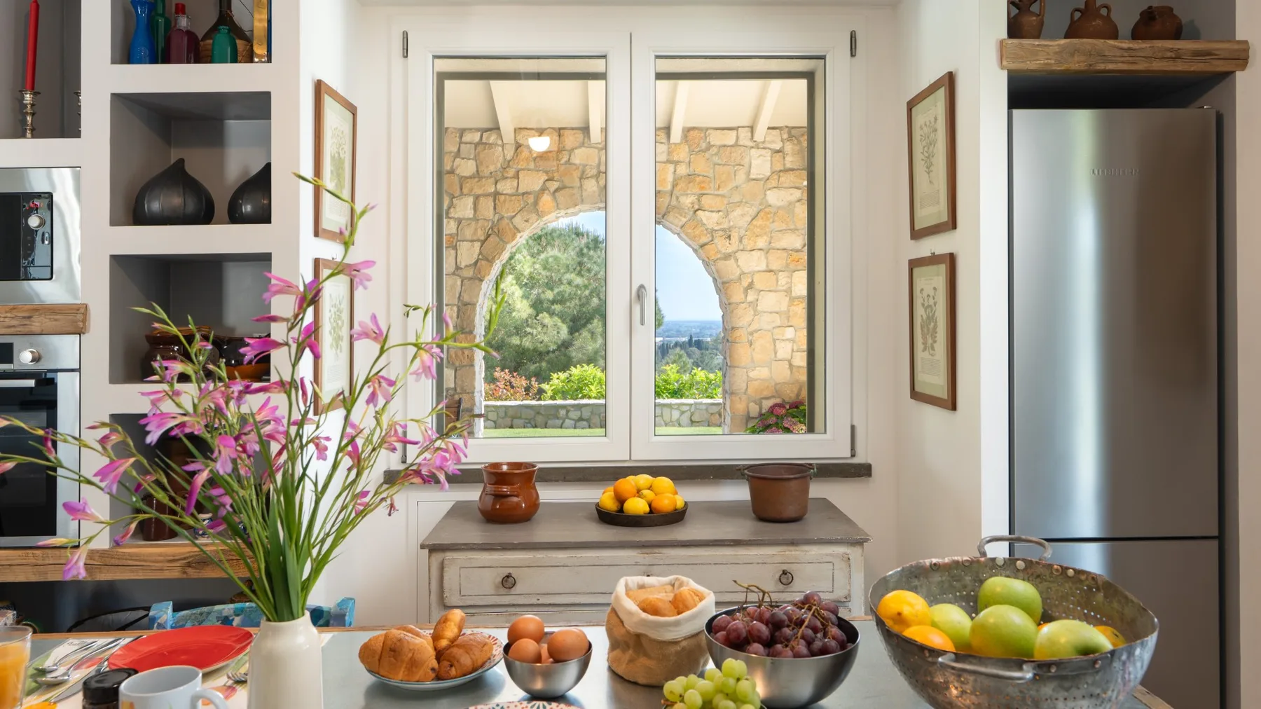 A bright kitchen with a counter of fruit and flowers beside a window.