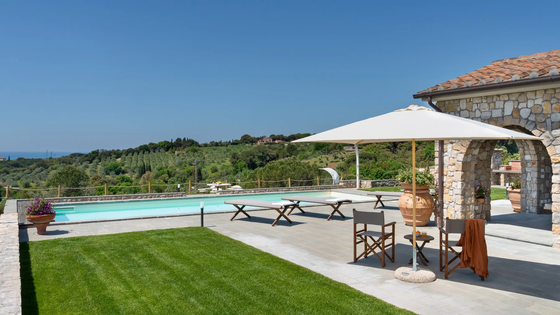 A sunny outdoor pool area with a lawn, seating, and a large umbrella.