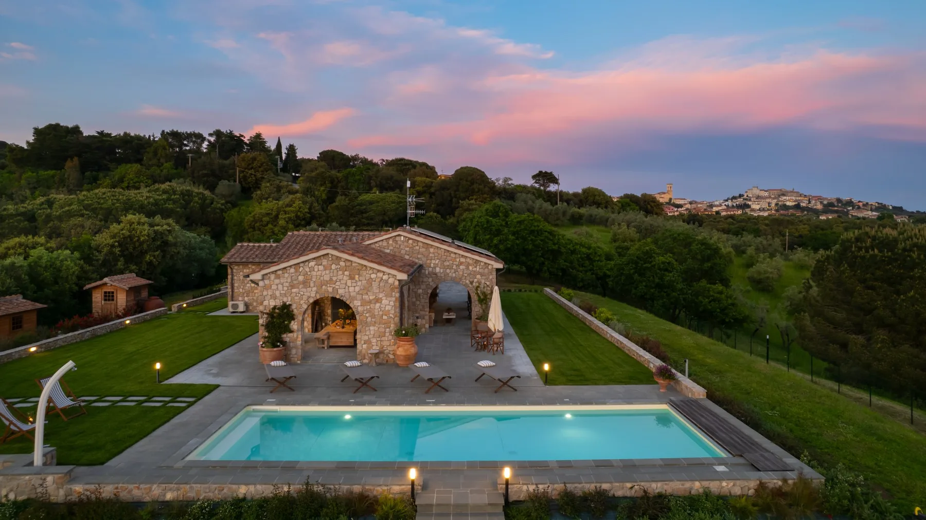 A stone villa and outdoor swimming pool sit amid green gardens beneath a colorful evening sky.