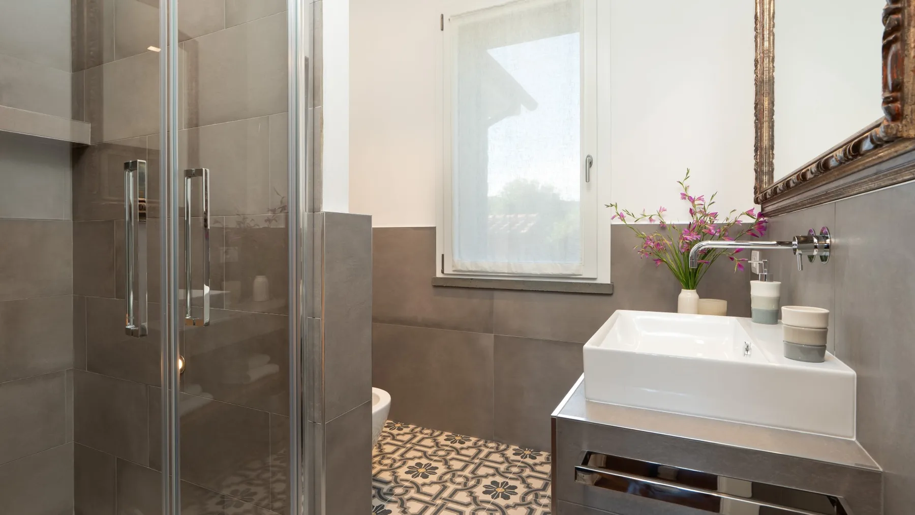 A bathroom with a glass shower, patterned floor, and sink beneath a window.
