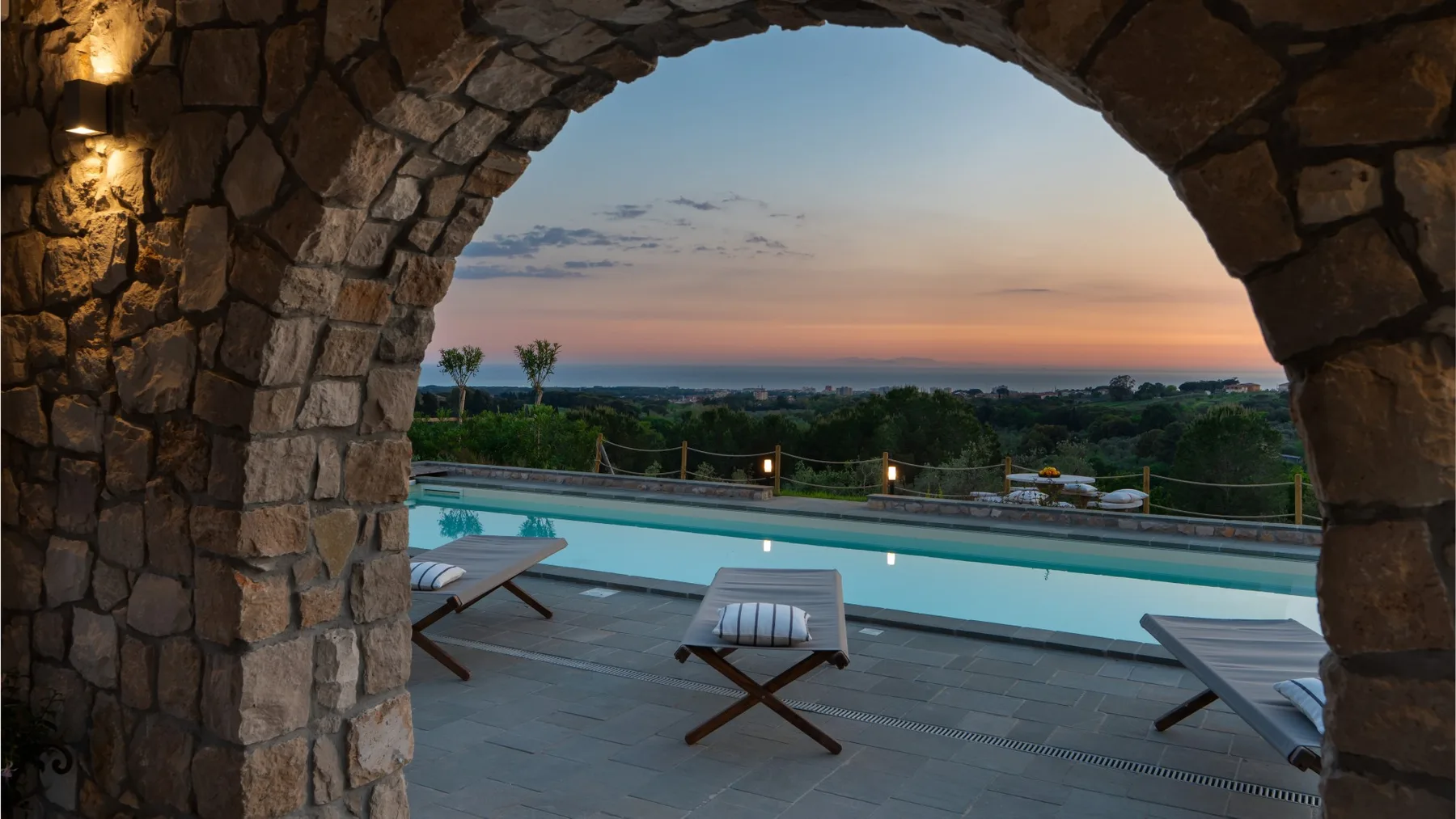 A swimming pool and loungers are framed by a stone arch at sunset.