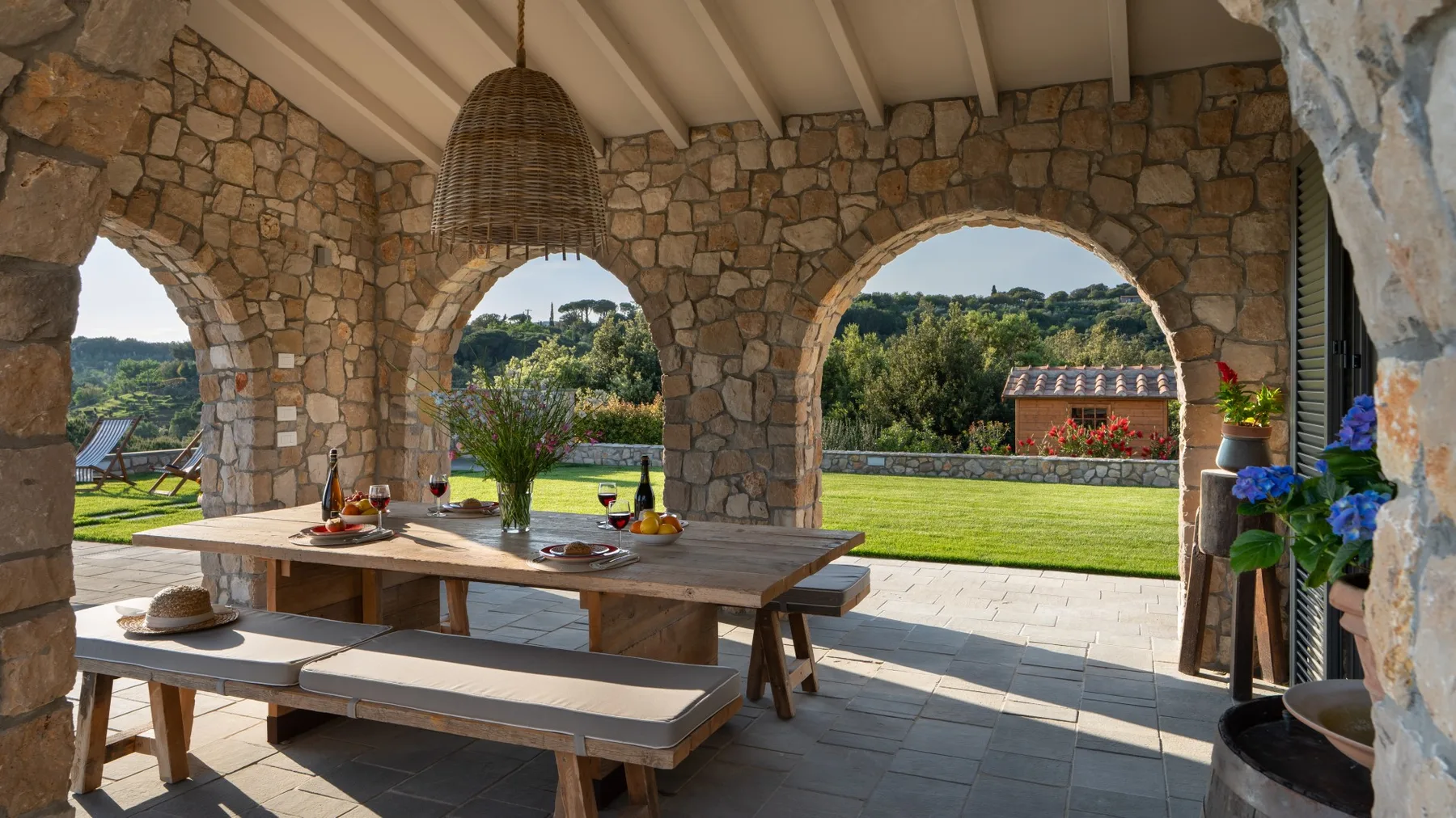 A stone dining area opens through arches onto a garden, with a long wooden table.