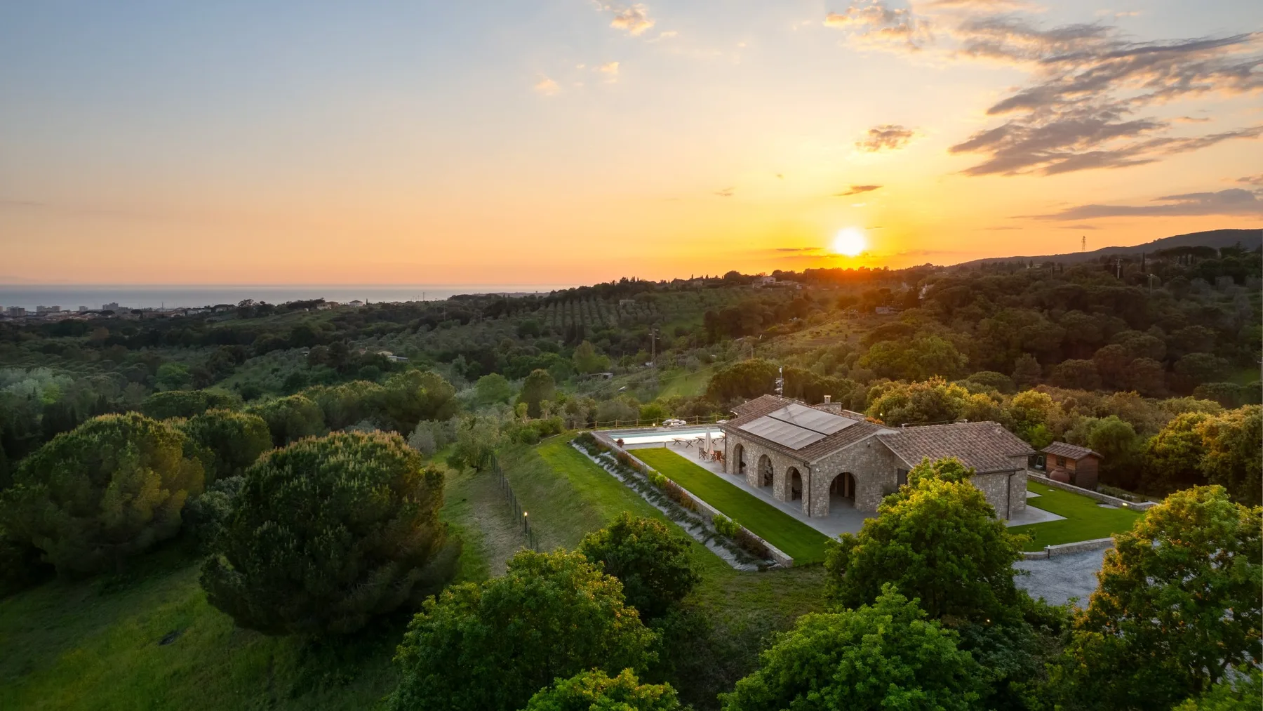 A countryside property sits among green trees beneath a colorful sunset sky.