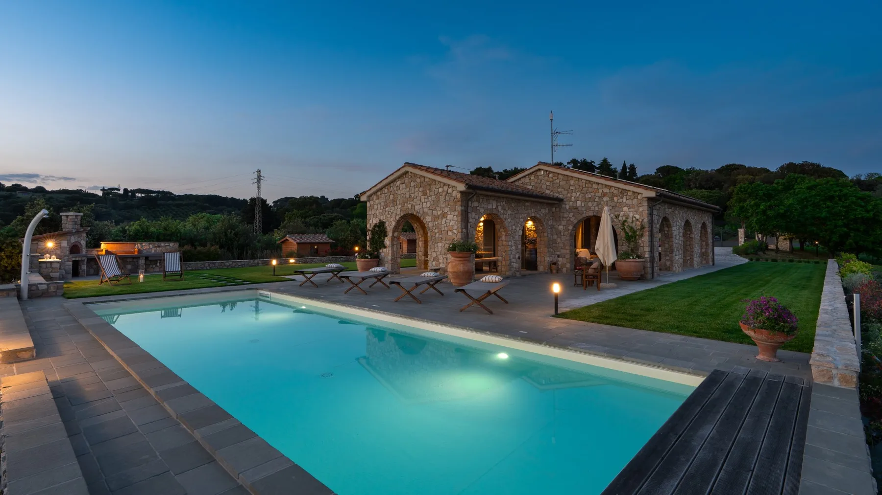 A swimming pool beside a stone building, with outdoor seating and greenery at dusk.