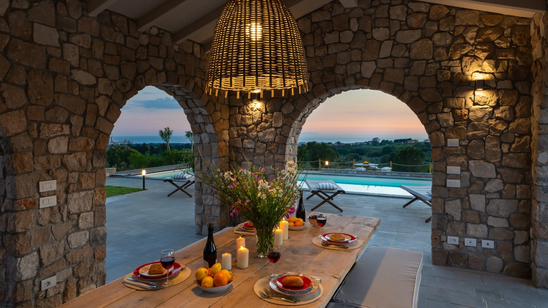 A stone dining area opens through arches onto a poolside terrace at dusk.
