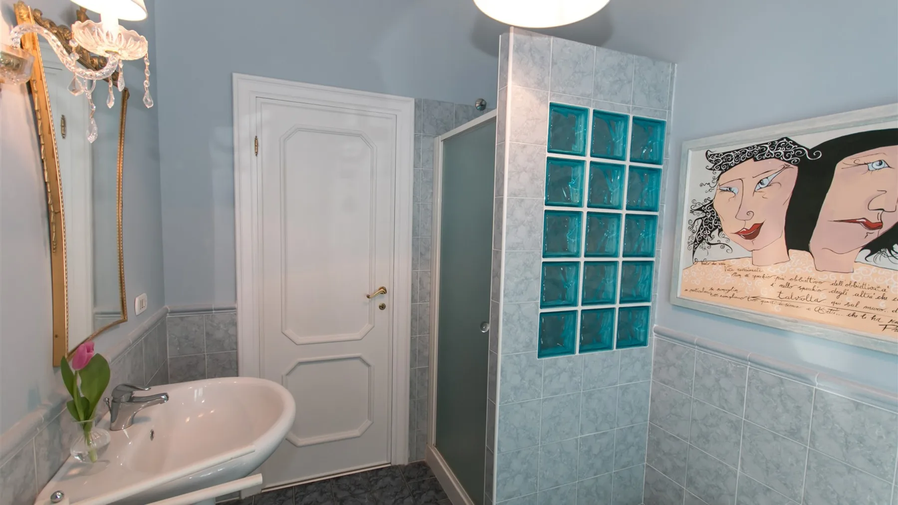 Un baño alicatado con lavabo blanco, puerta con paneles, separador de bloques de vidrio turquesa y arte enmarcado.