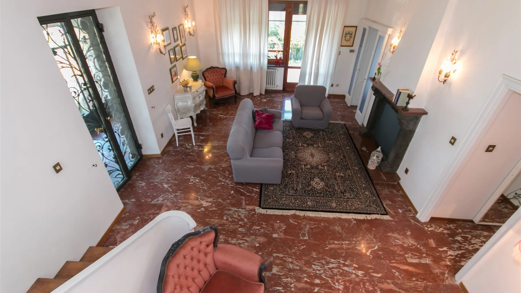 Una sala luminosa tiene sofás grises, una alfombra estampada y un suelo de mármol bajo ventanas altas.