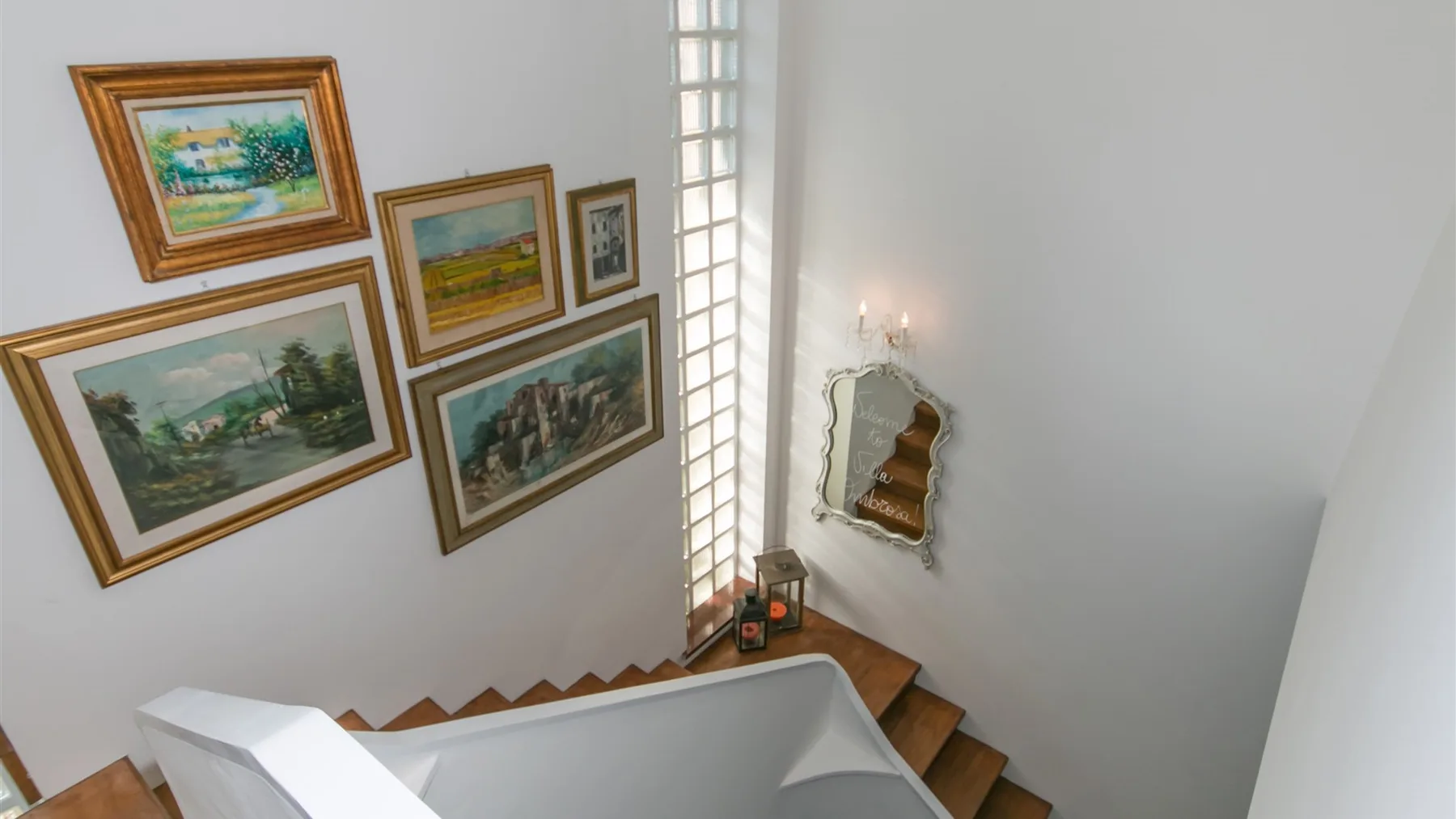 Una escalera blanca desciende junto a cuadros de paisajes, con una ventana alta y un pequeño espejo en la pared.