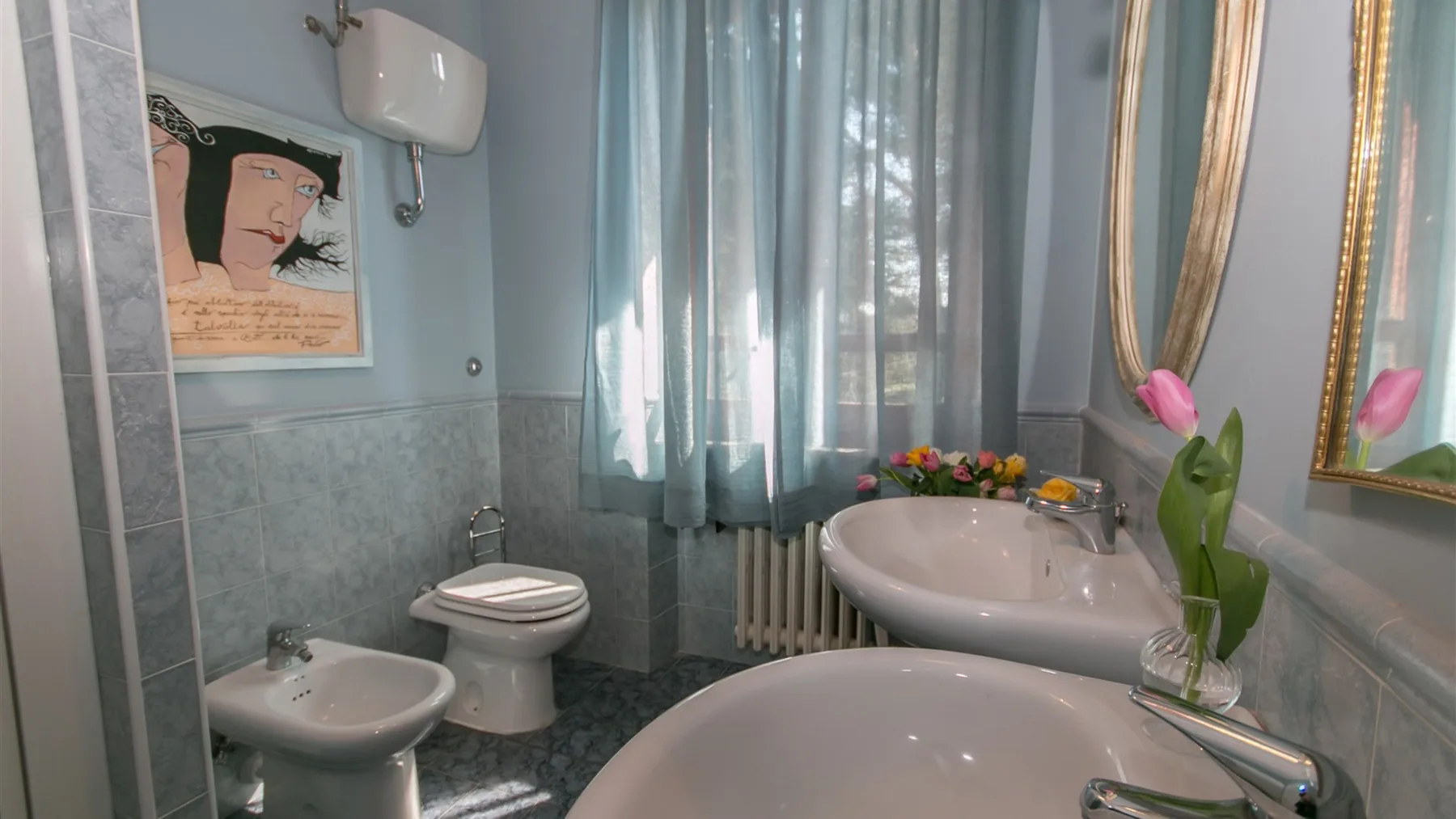 Un baño alicatado tiene bañera, bidé, inodoro y lavabo bajo cortinas azules translúcidas.