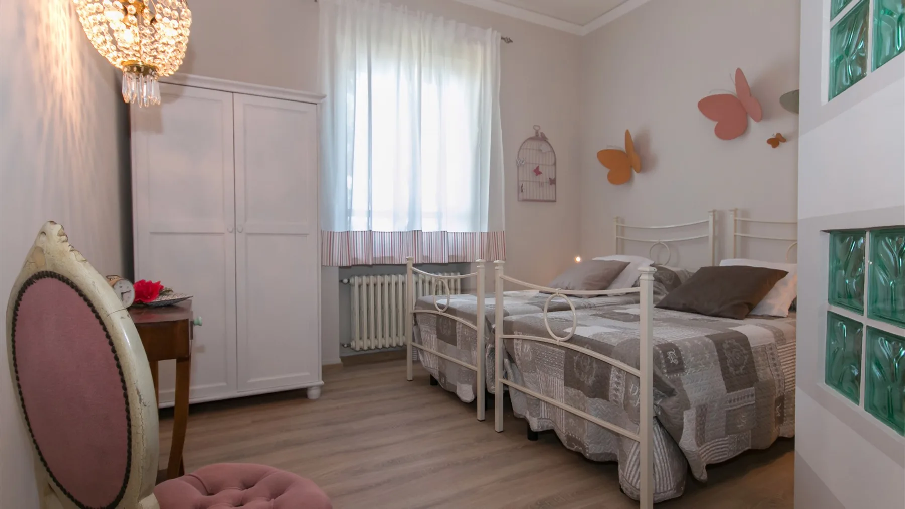 Un dormitorio luminoso tiene dos camas individuales, un armario blanco y una gran ventana con cortinas.