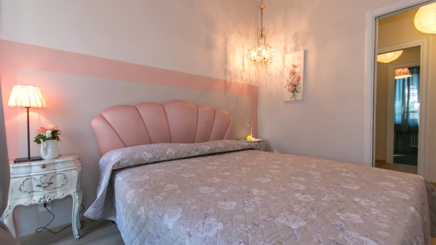 Un dormitorio con una cama grande, una mesita blanca tallada y una lámpara de araña encendida.
