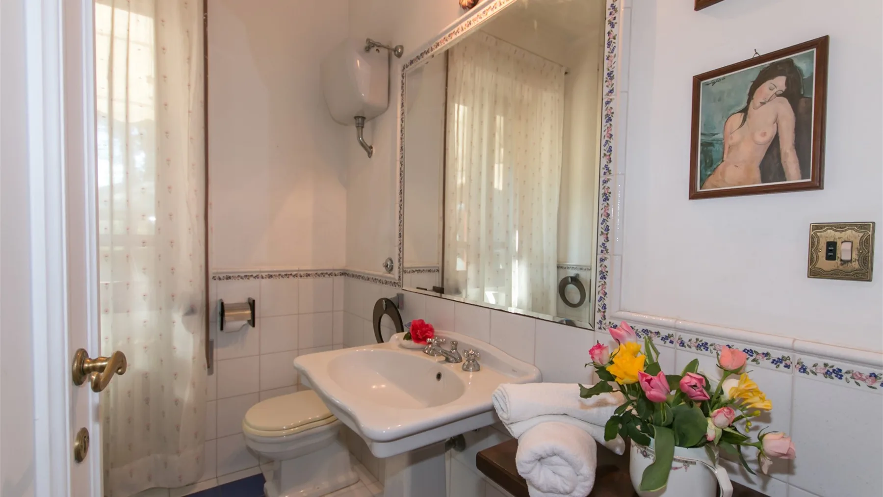 Un baño alicatado con lavabo blanco, espejo grande, inodoro y flores junto al lavabo.