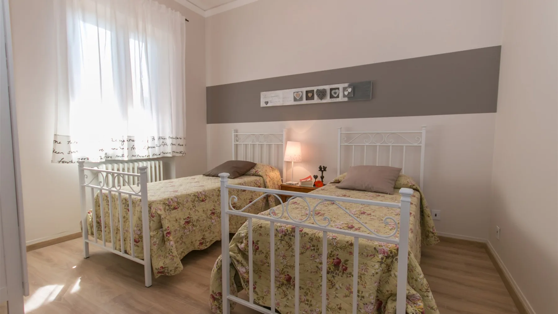 Un dormitorio luminoso con dos camas de metal blanco, ropa de cama floral y una ventana alta con cortinas translúcidas.
