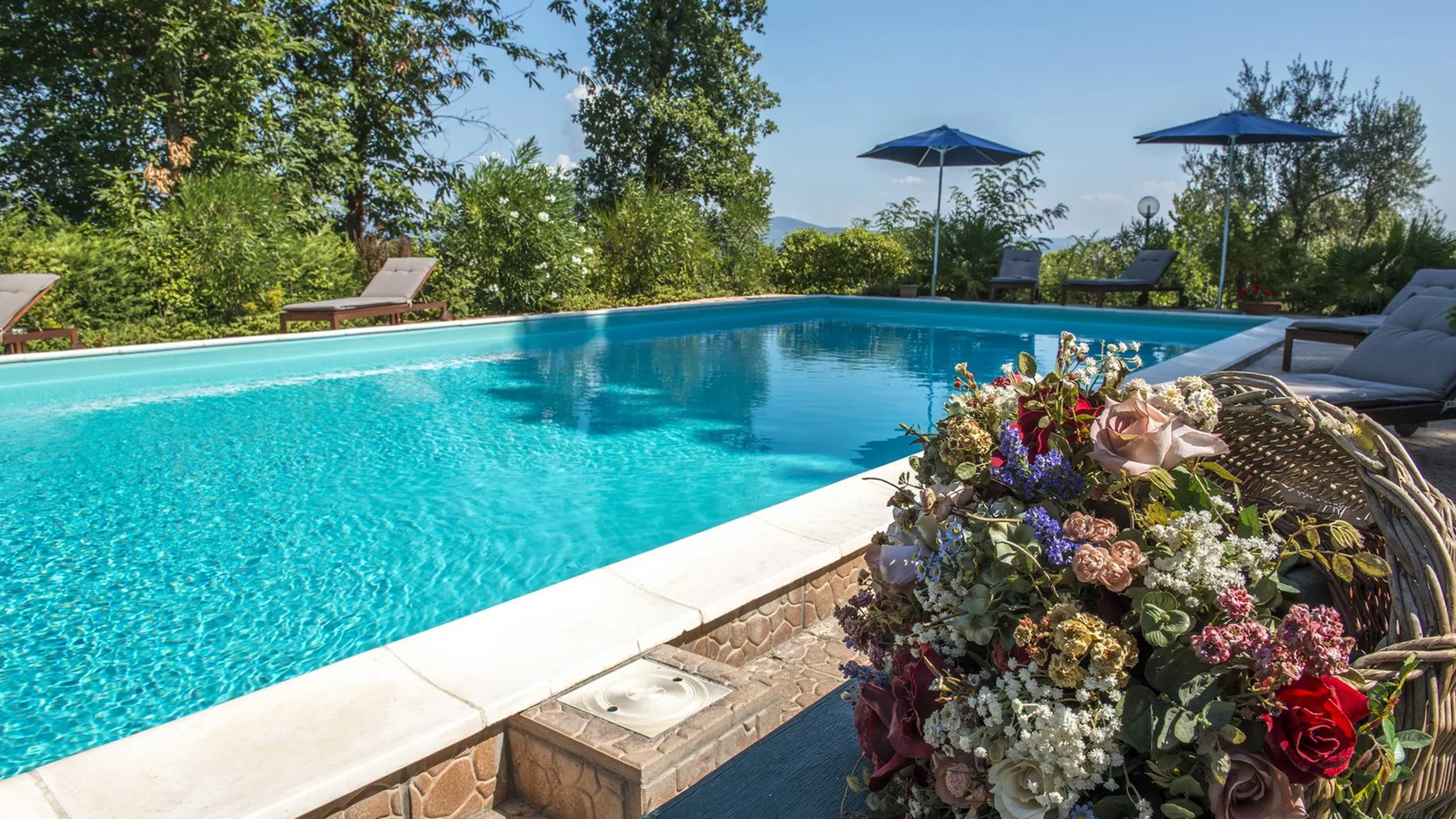 Una piscina turquesa está rodeada de pavimento de piedra, tumbonas, sombrillas azules y un colorido arreglo floral.