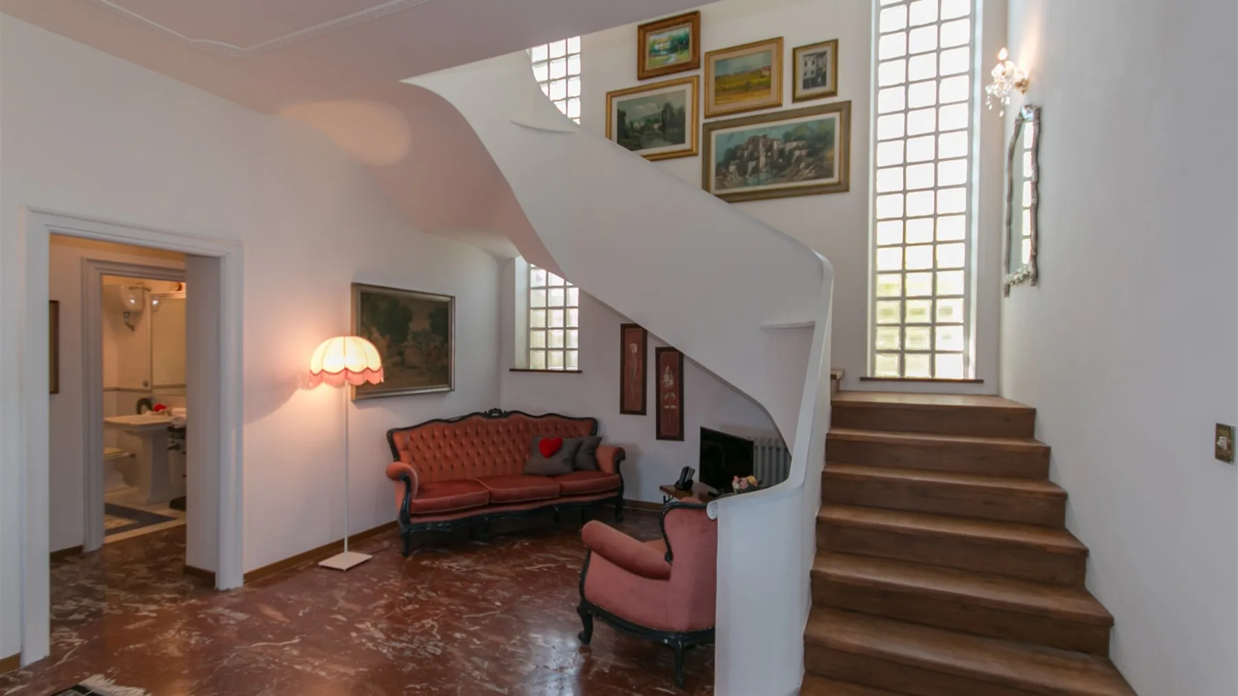 Una escalera curva se eleva sobre una zona de estar con sofá rojo, sillón, cuadros y ventanas altas.
