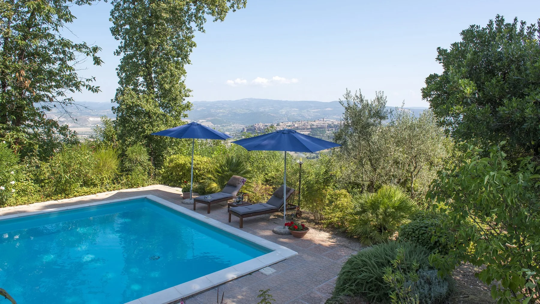 Una piscina al aire libre con sombrillas azules y tumbonas da a un valle verde.