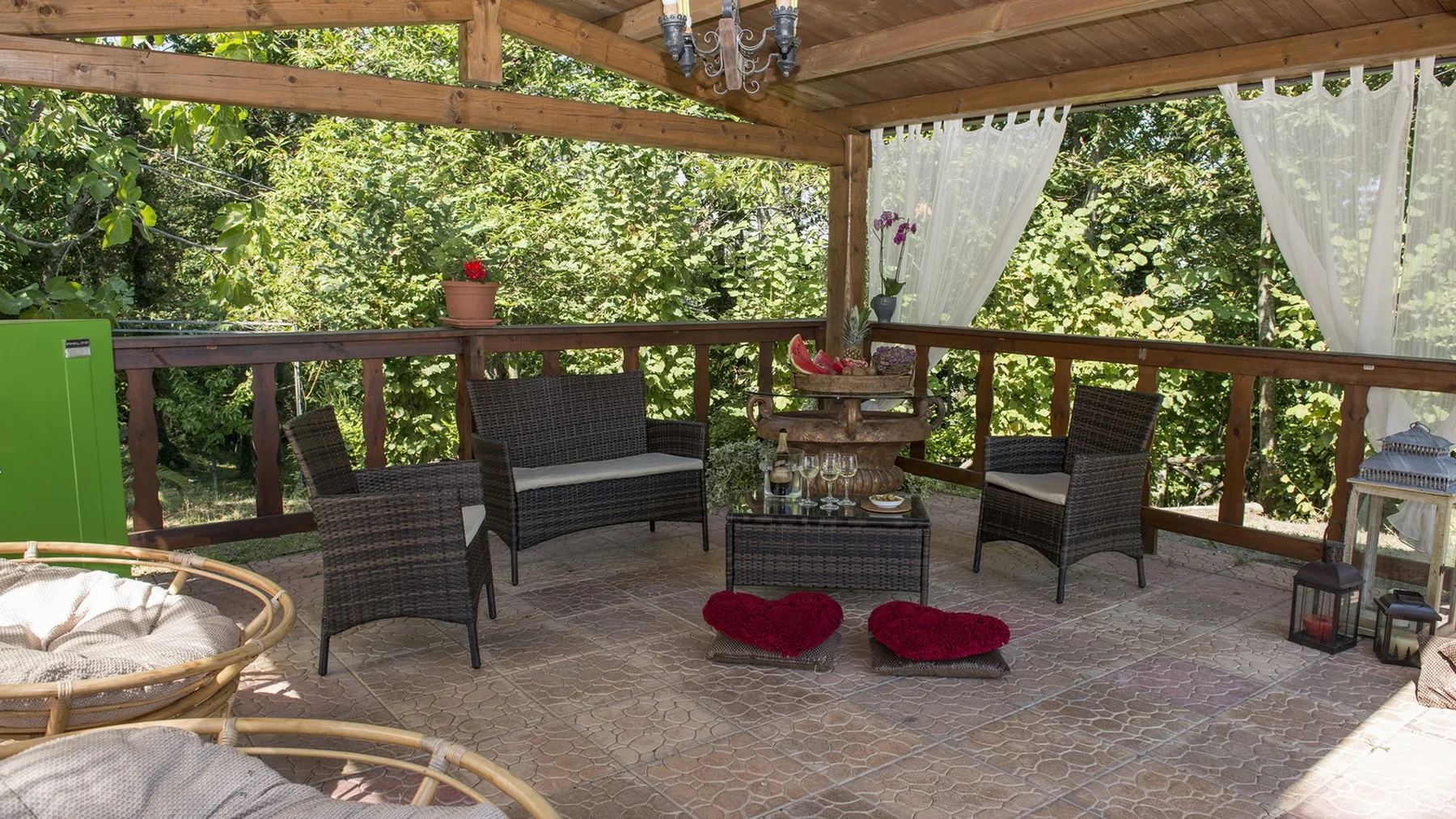 Un patio cubierto de madera tiene asientos de mimbre alrededor de una mesa con tapa de vidrio, con árboles al otro lado de la barandilla.