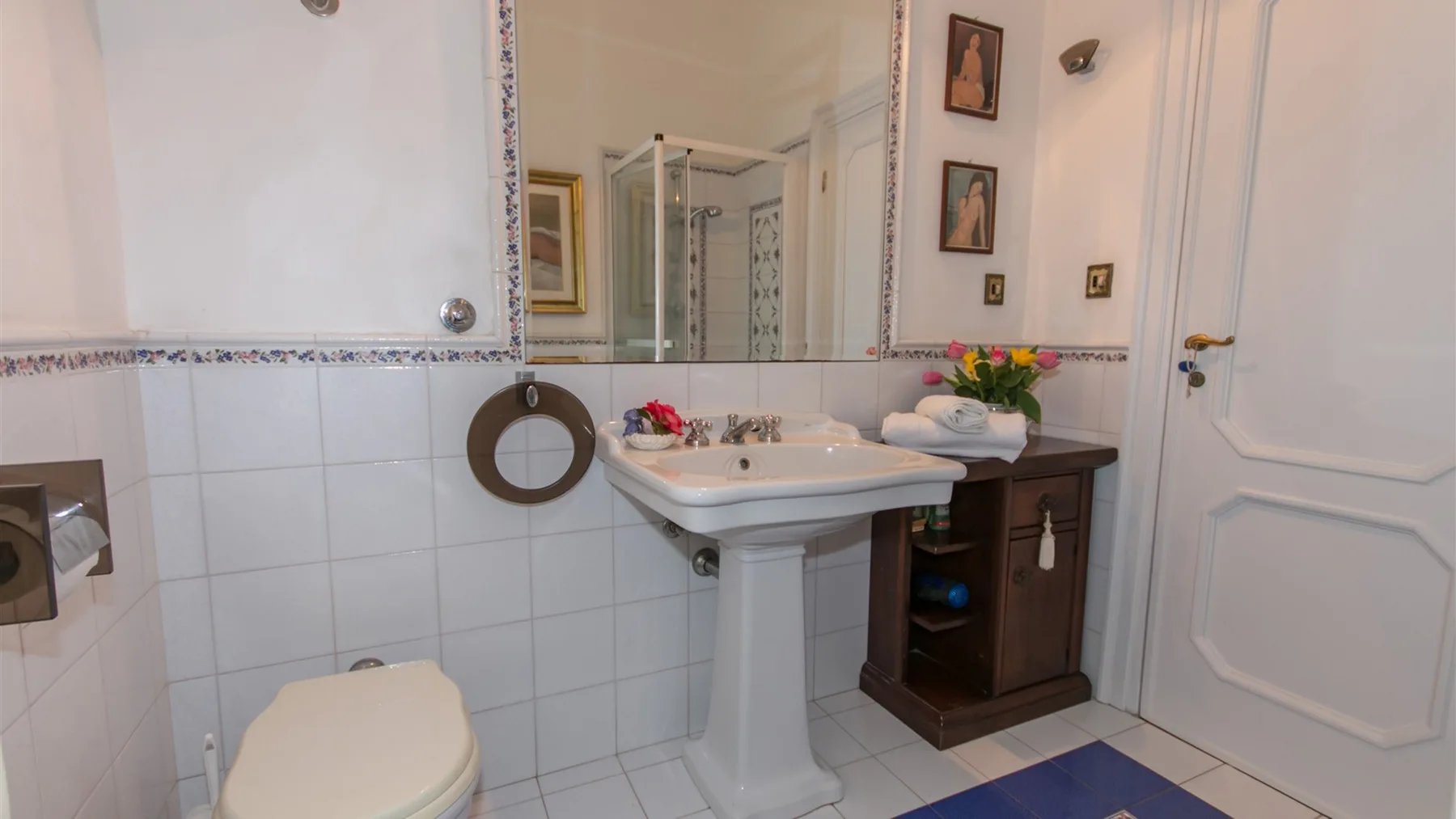 Un baño alicatado tiene un lavabo con pedestal, un inodoro, un mueble de madera y una alfombrilla azul.