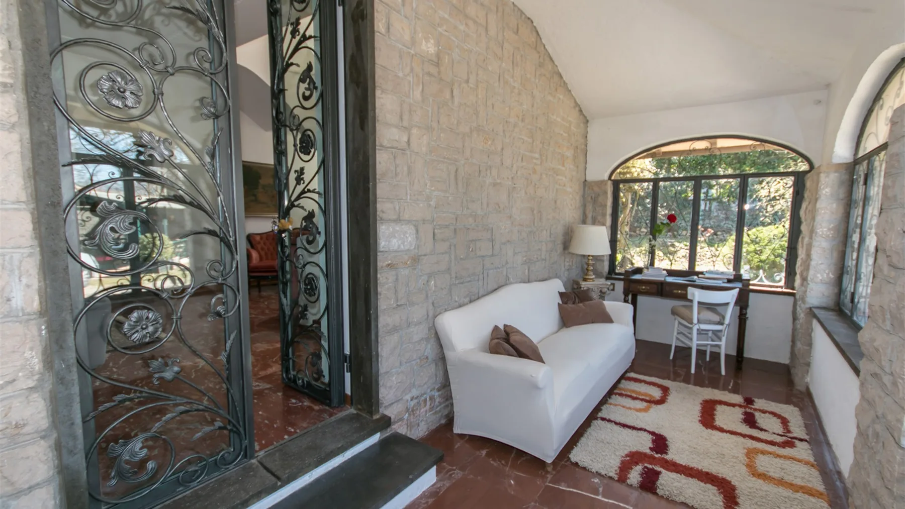 Una habitación de paredes de piedra tiene un sofá blanco, una alfombra estampada y una mesa junto a grandes ventanas arqueadas.
