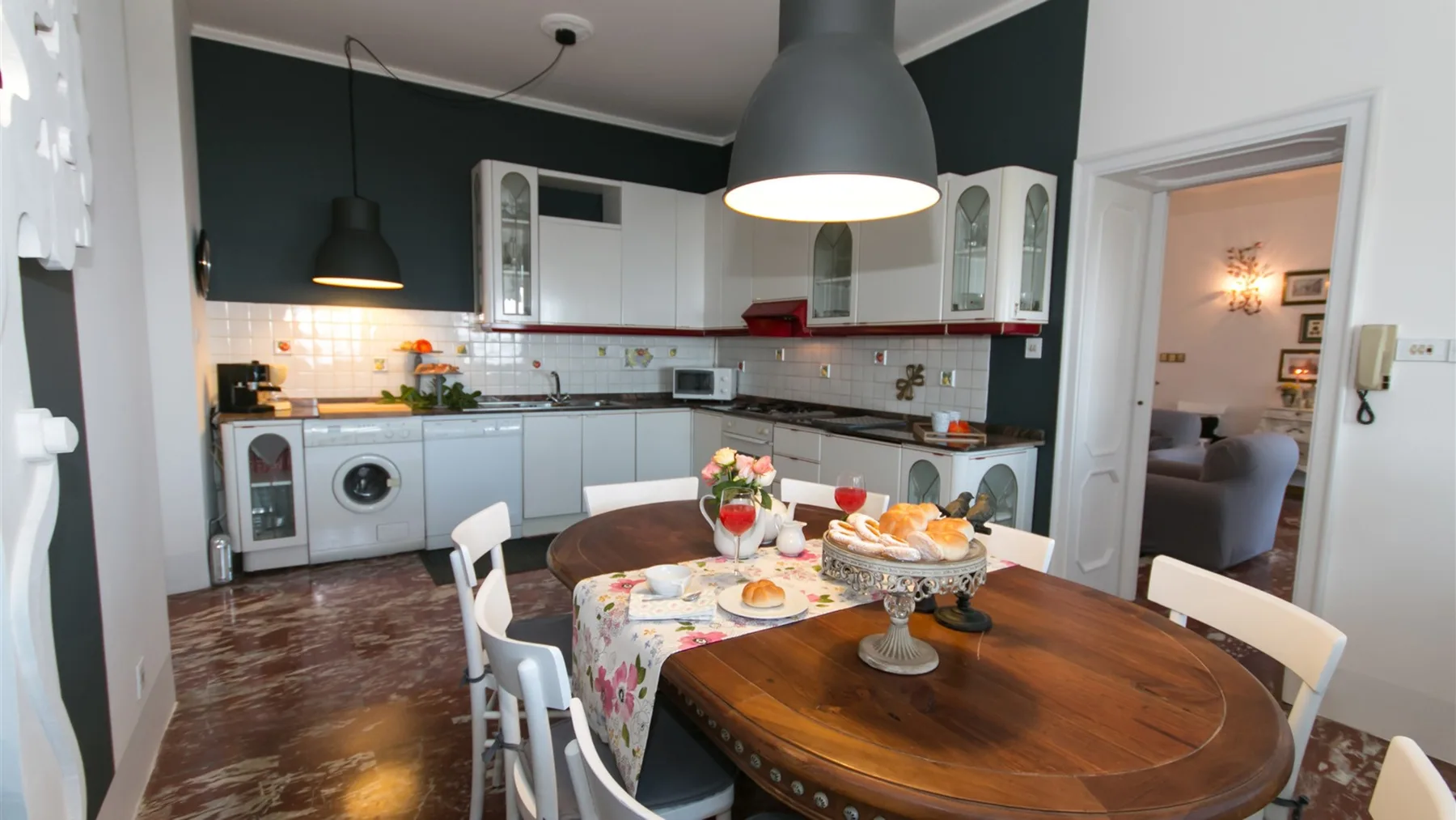 Un comedor con una mesa de madera servida con comida, junto a una cocina con armarios blancos y electrodomésticos.