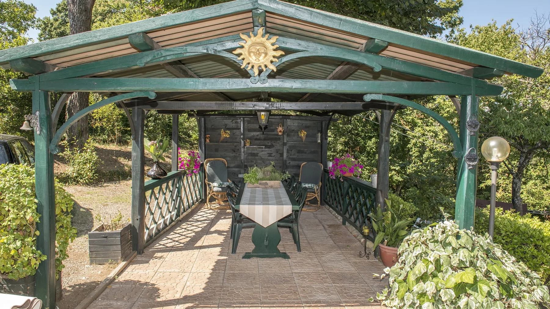 Un comedor cubierto en el jardín tiene una mesa larga, sillas, barandillas decorativas y plantas con flores.