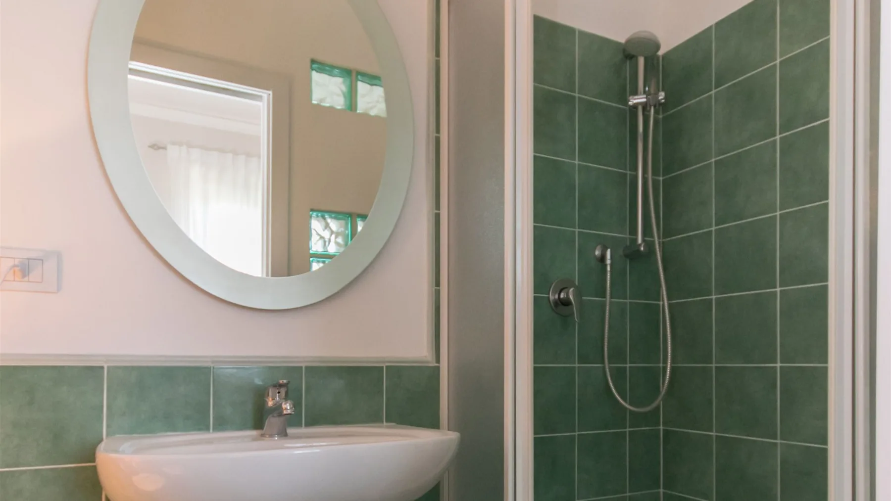 Un baño cuenta con un lavabo blanco bajo un gran espejo redondo y una ducha con paredes de azulejos verde oscuro.