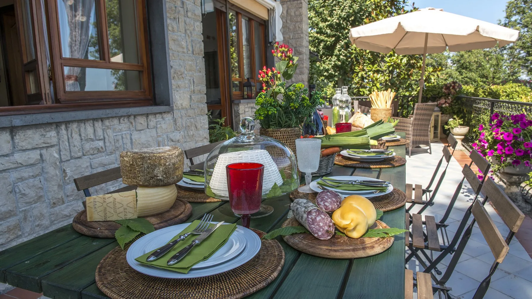 Una mesa de comedor en un patio junto a una casa de piedra está preparada con platos y comida bajo una gran sombrilla.