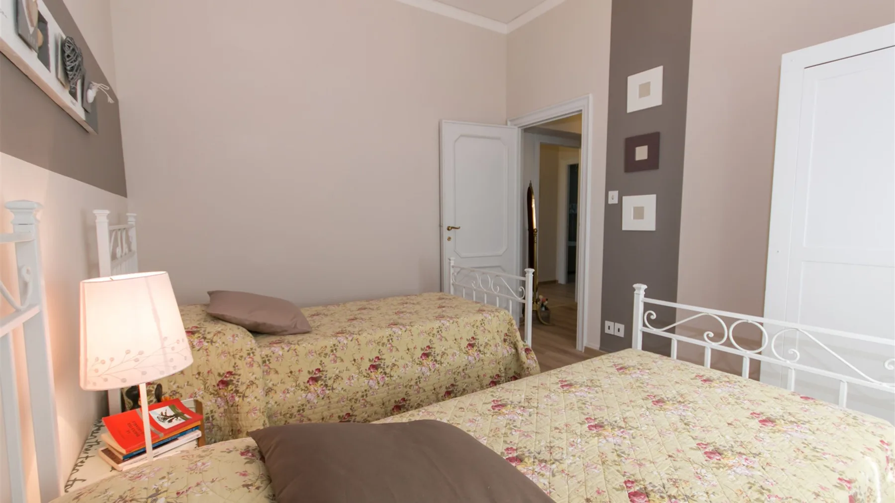 Un dormitorio con dos camas cubiertas con colchas florales, cabeceros blancos de metal y una lámpara de mesita.
