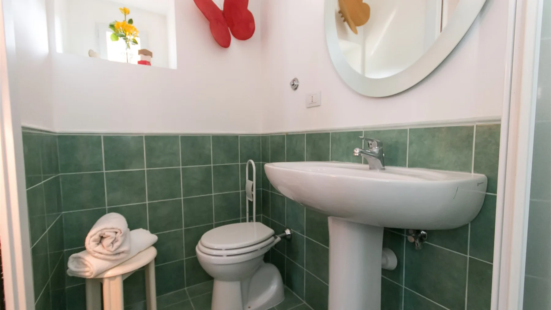 Un baño pequeño tiene paredes de azulejos verdes, un lavabo con pedestal y un inodoro junto a un taburete con toallas dobladas.