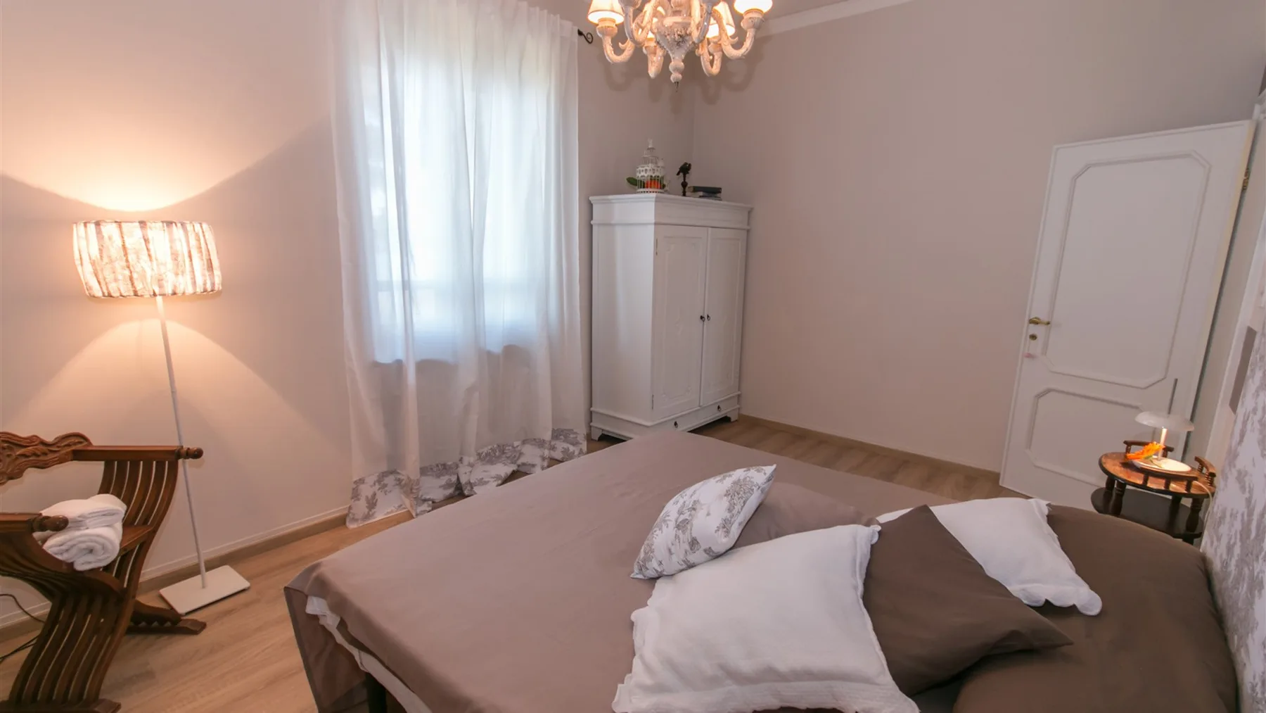 Un dormitorio con cama doble, ventana con cortinas translúcidas, armario blanco y lámpara de araña.