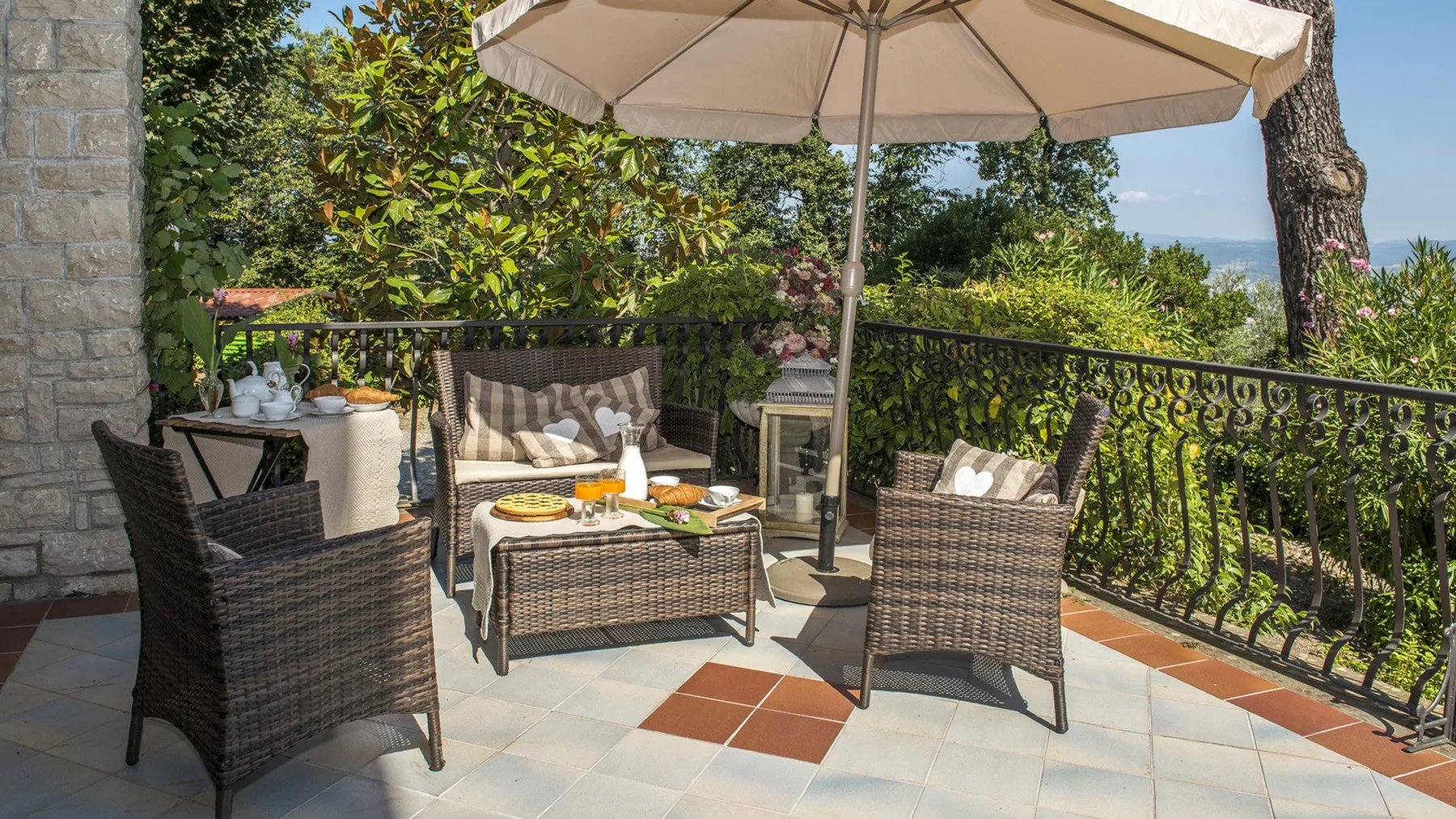 Una terraza embaldosada tiene asientos de mimbre y una mesa de centro bajo una gran sombrilla, con plantas al otro lado de la barandilla.