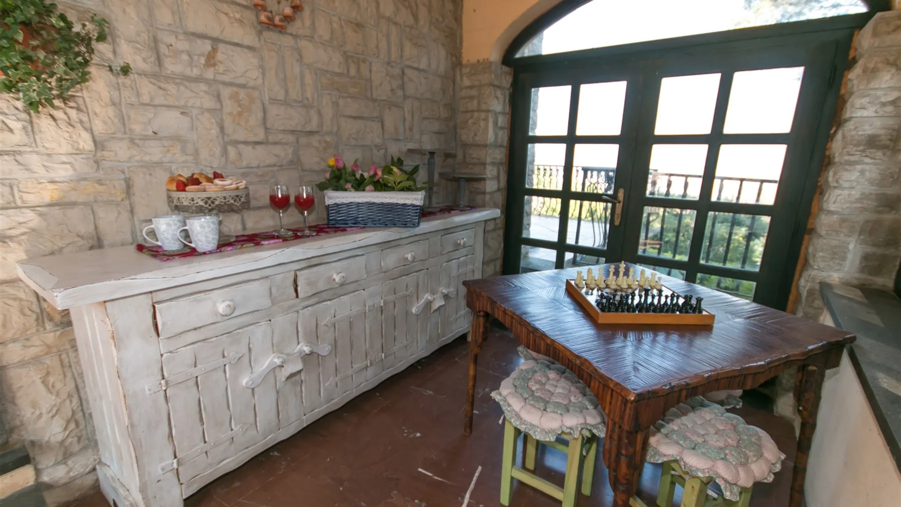 Una estancia con paredes de piedra tiene un aparador blanco, una mesa de ajedrez y grandes puertas acristaladas.