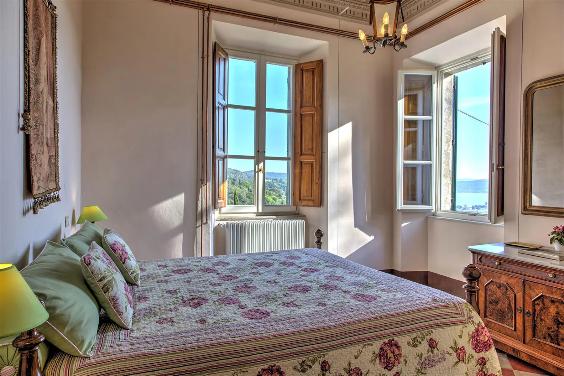 Un dormitorio con colcha floral, ventanas abiertas y una cómoda de madera iluminada por la luz del día.