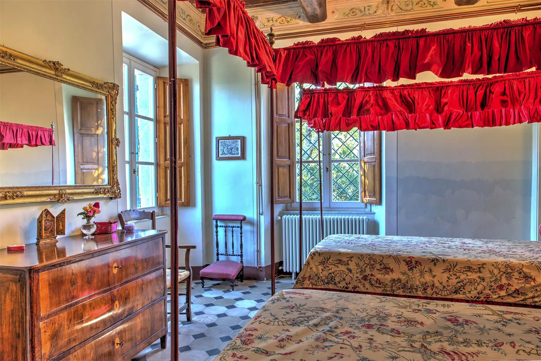 Un dormitorio con dos camas estampadas, cortinajes rojos, una cómoda de madera y altas ventanas emplomadas.