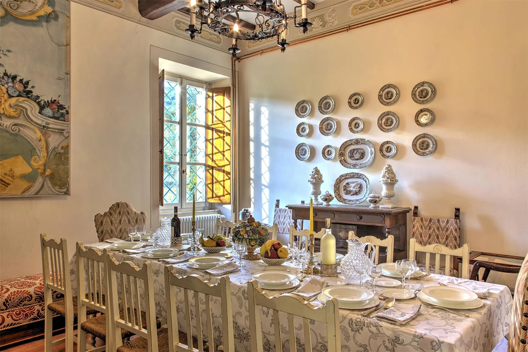 Un comedor ornamentado presenta una mesa larga puesta, platos decorativos y altas ventanas de vidrio emplomado.