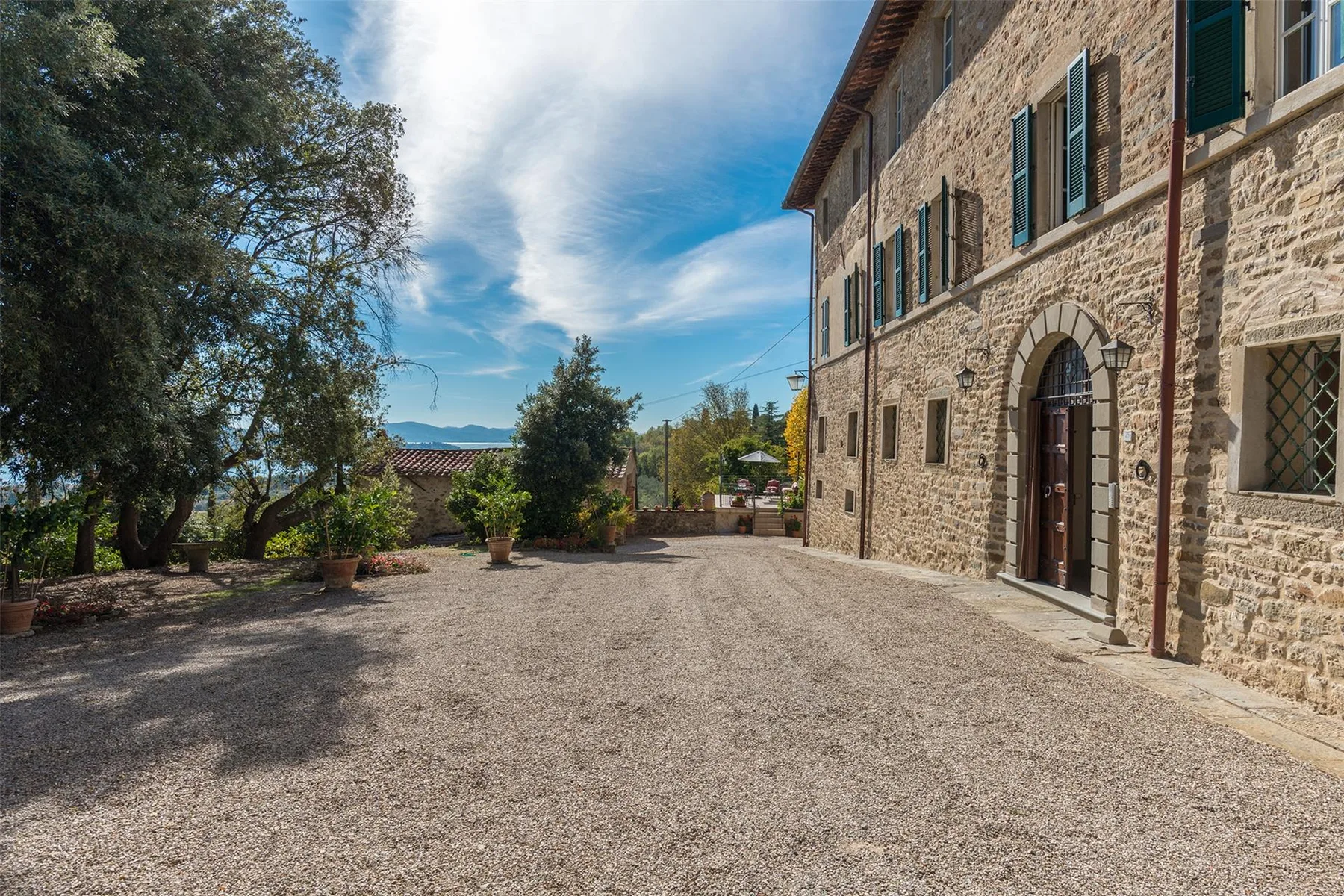 Un patio de grava junto a un edificio de piedra, con puertas arqueadas, ventanas con contraventanas y árboles.