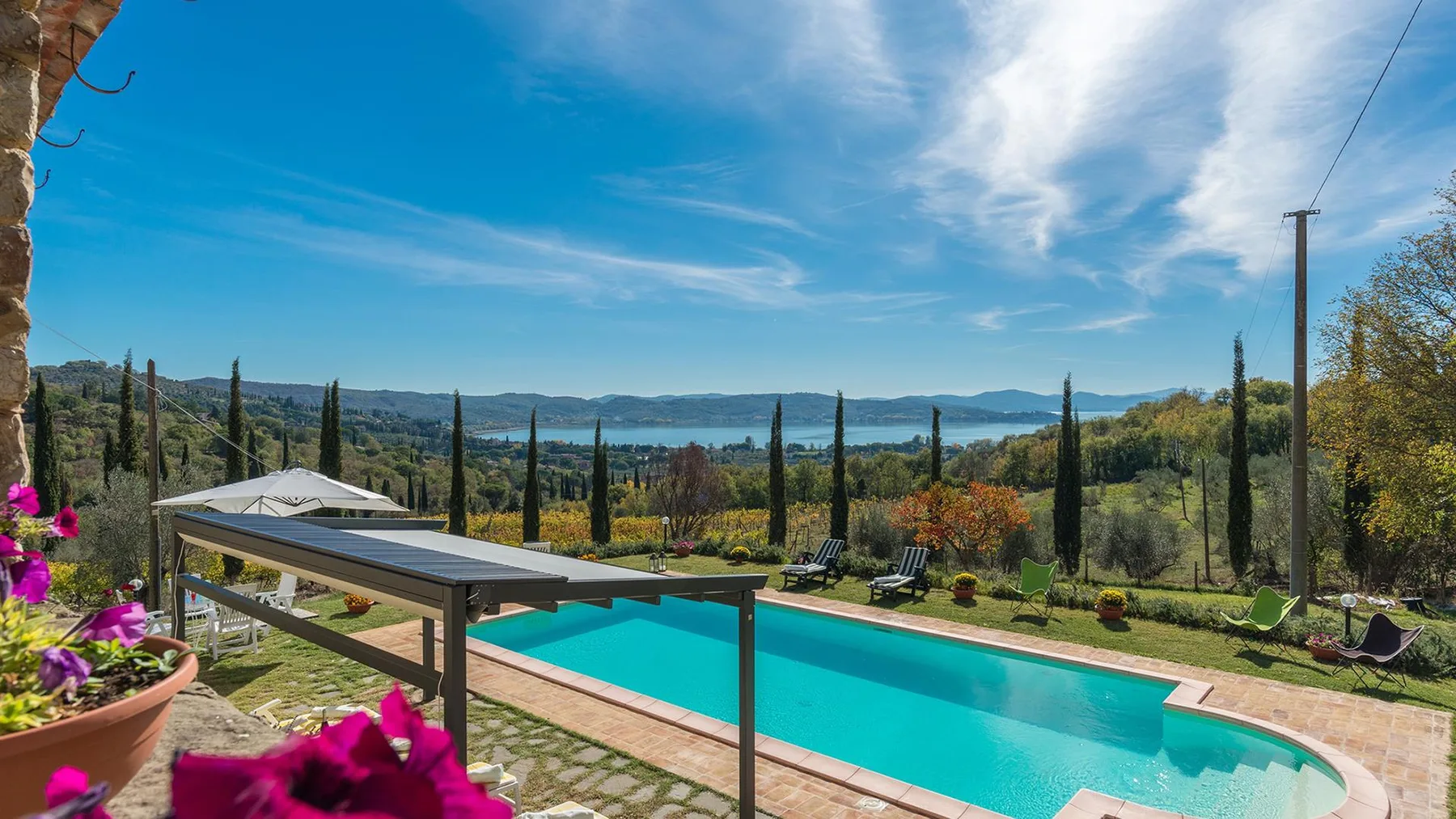 Una piscina turquesa da a un jardín ajardinado, cipreses y un lago a lo lejos.