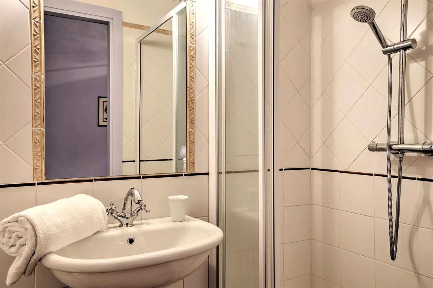 Un baño alicatado tiene un lavabo blanco con una toalla enrollada junto a una ducha con mampara de vidrio.
