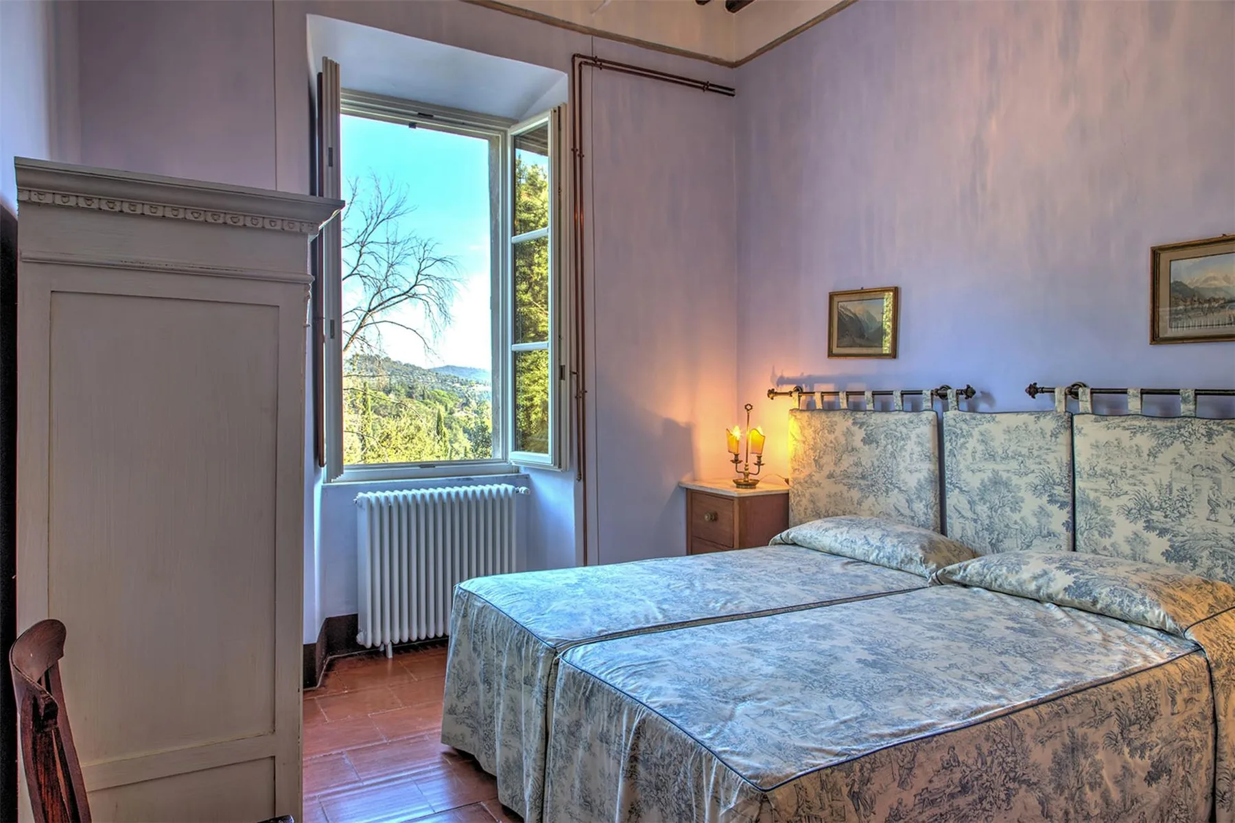 Un dormitorio con dos camas, ropa de cama estampada en tonos azul grisáceo y una ventana con vistas a colinas verdes.