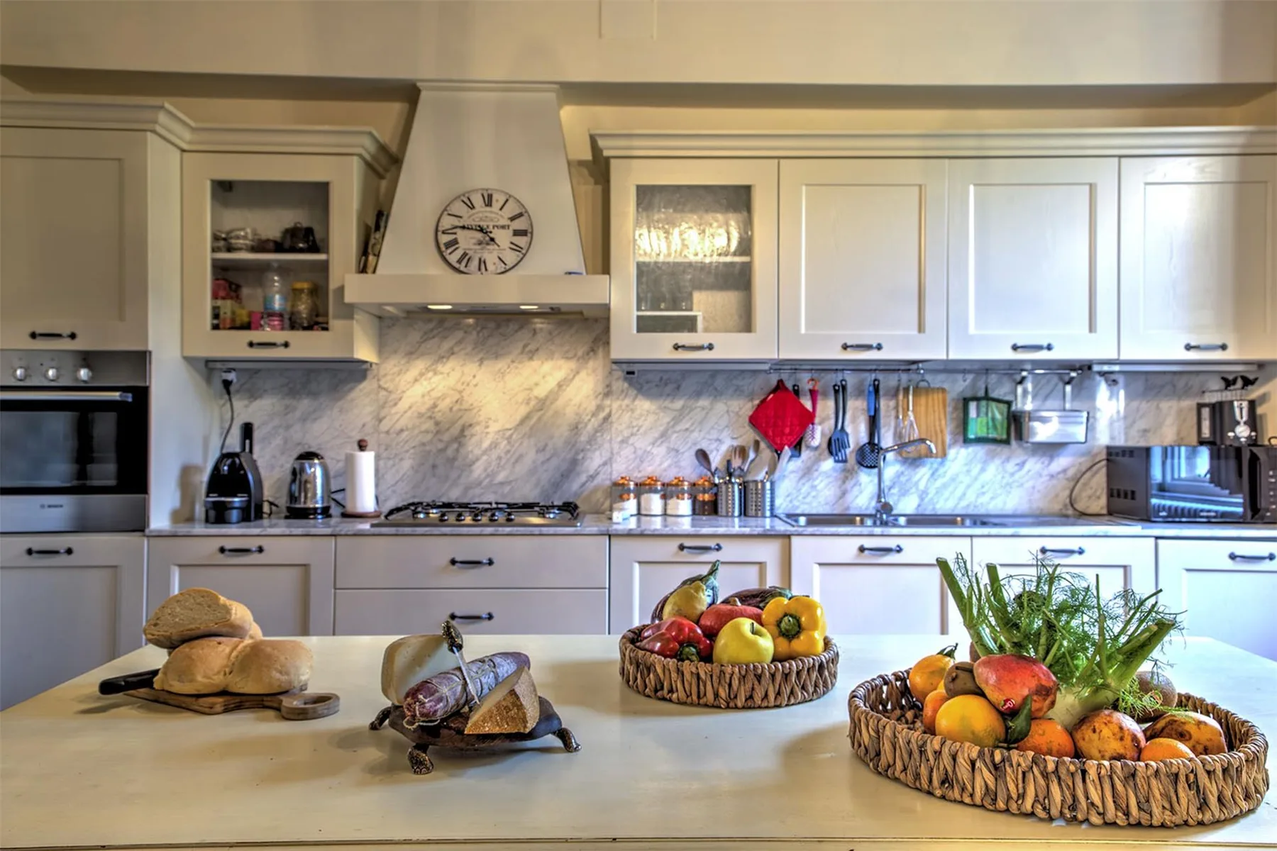 Una cocina luminosa con gabinetes blancos, un salpicadero de mármol y cestas de fruta en la isla.
