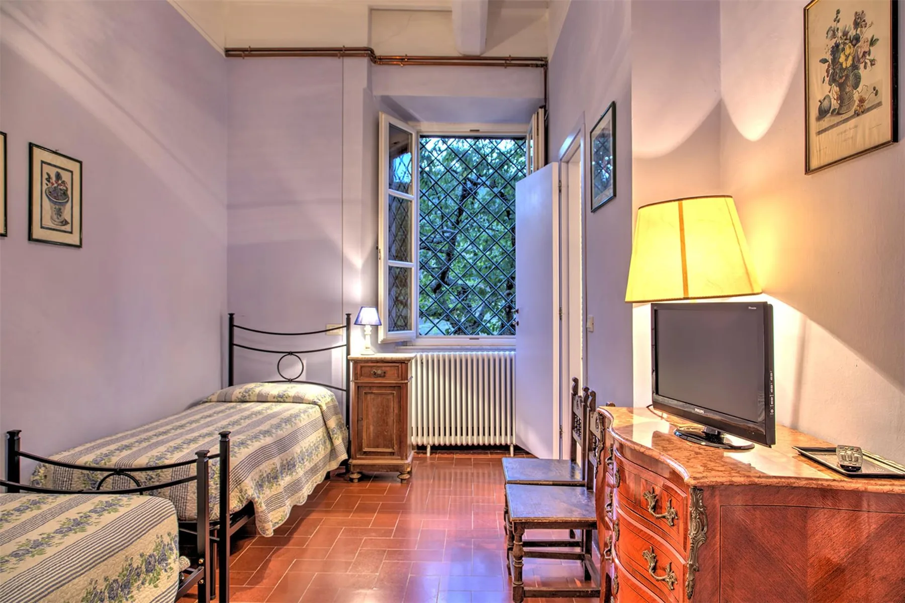 Un dormitorio amueblado tiene dos camas con estructura metálica, una cómoda de madera y un televisor junto a una ventana abierta.