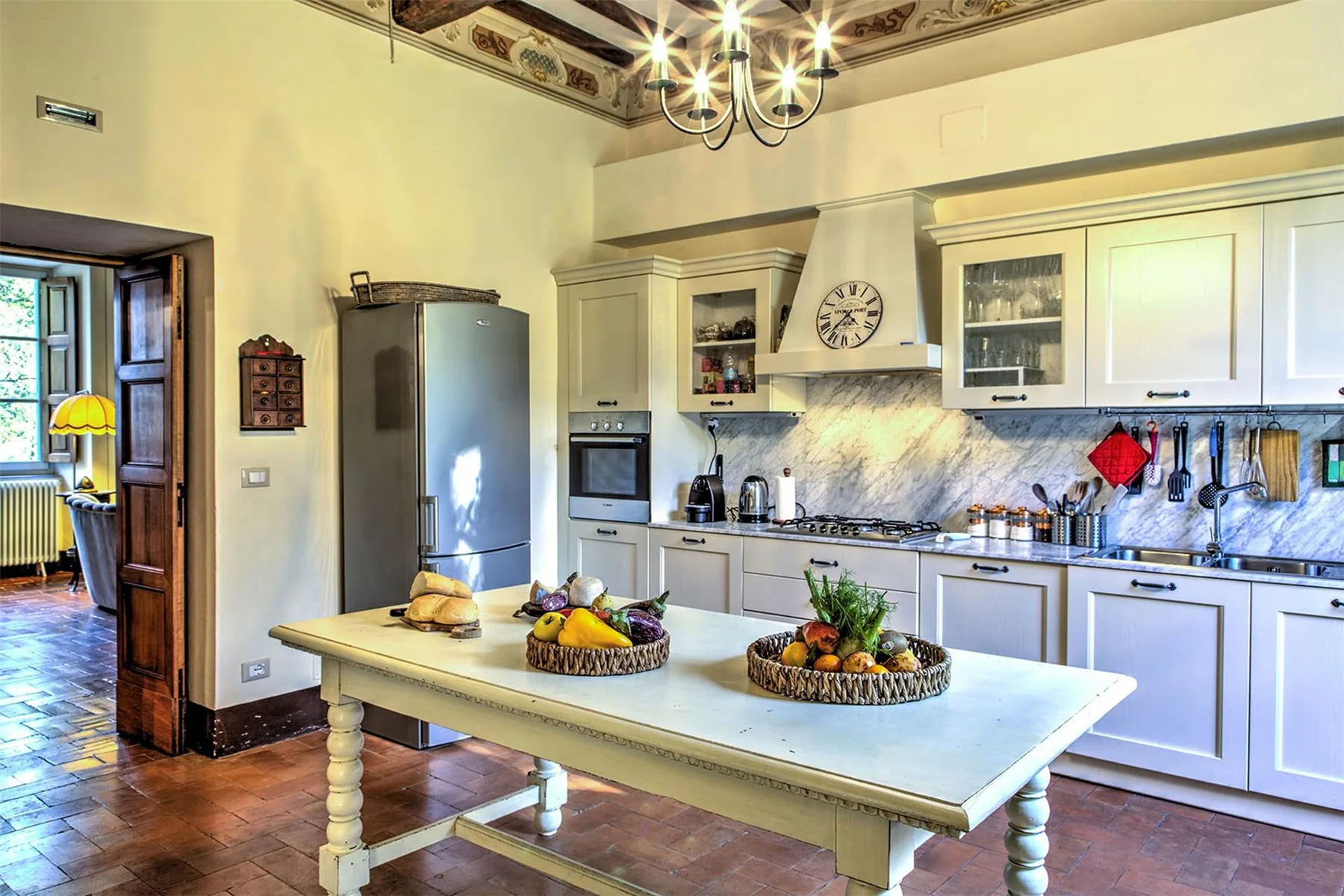 Una cocina luminosa cuenta con armarios blancos, una isla central con cestas de frutas y una lámpara bajo un techo pintado.